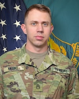 Man in U.S. Army uniform standing in front of two flags.