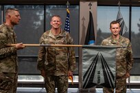 Two men in uniform stand at attention in front of a man in uniform holding a guidon