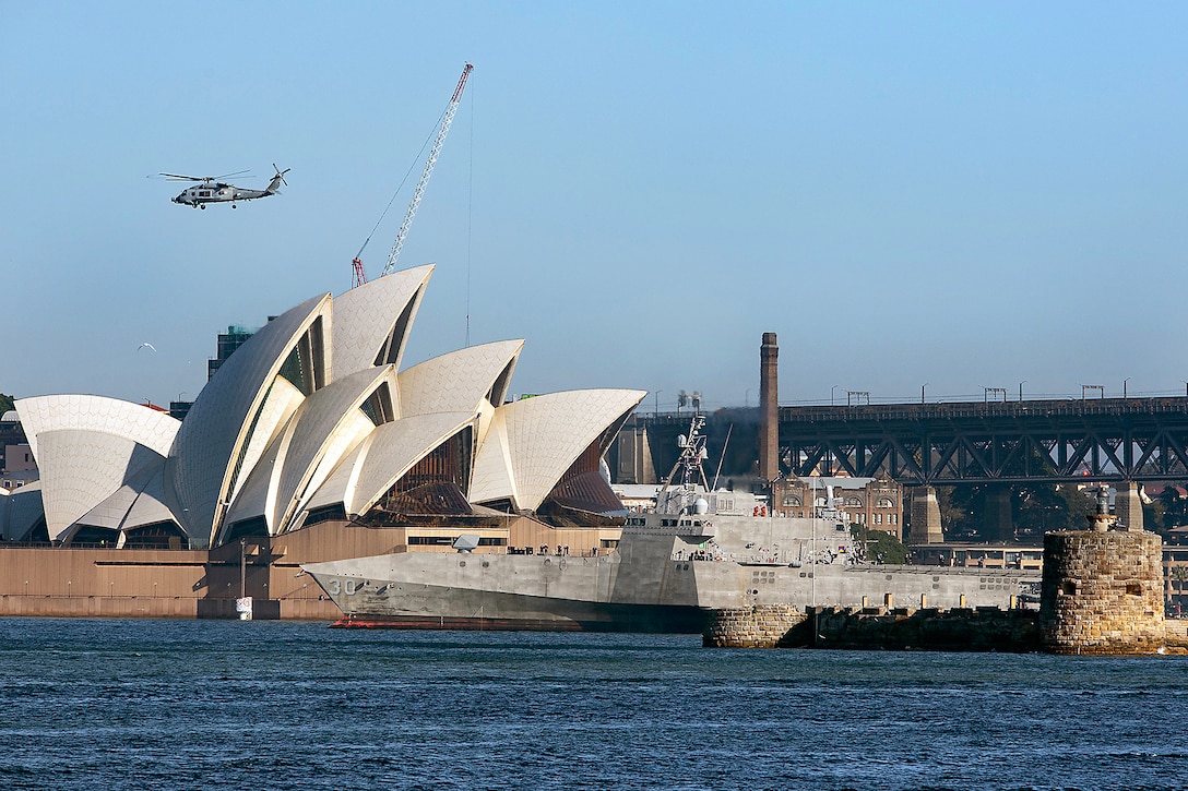 A ship sits in a harbor parallel to an opera house as a helicopter flies above with a bridge in the background.