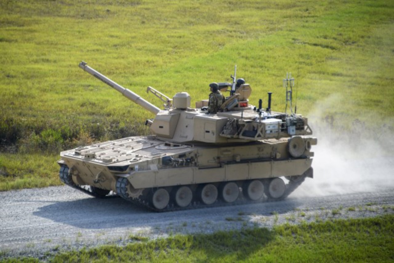 A service member looks out of the top of a tank moving down a dirt path.