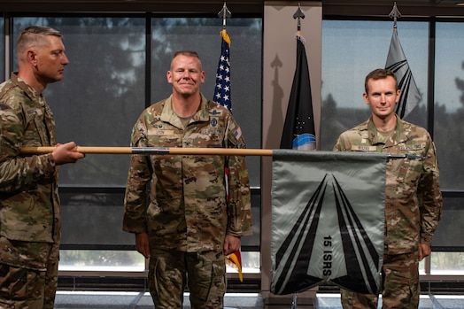  Two men in uniform stand at attention in front of a man in uniform holding a guidon