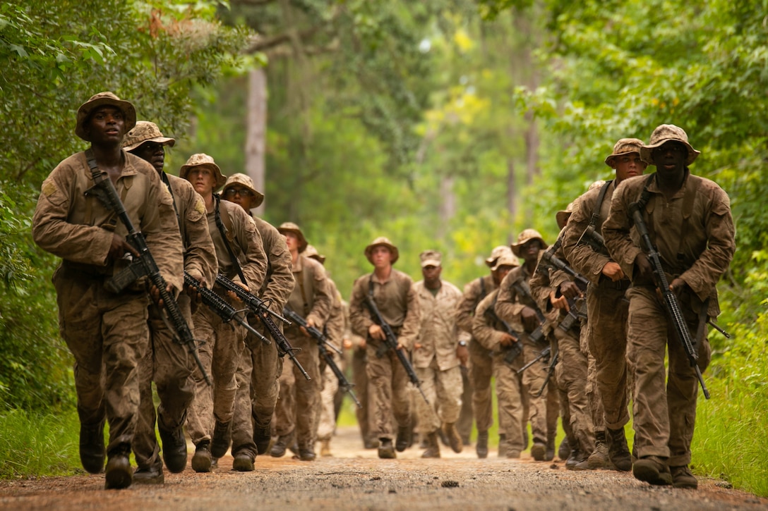 Marine carrying weapons march in two rows through the woods.