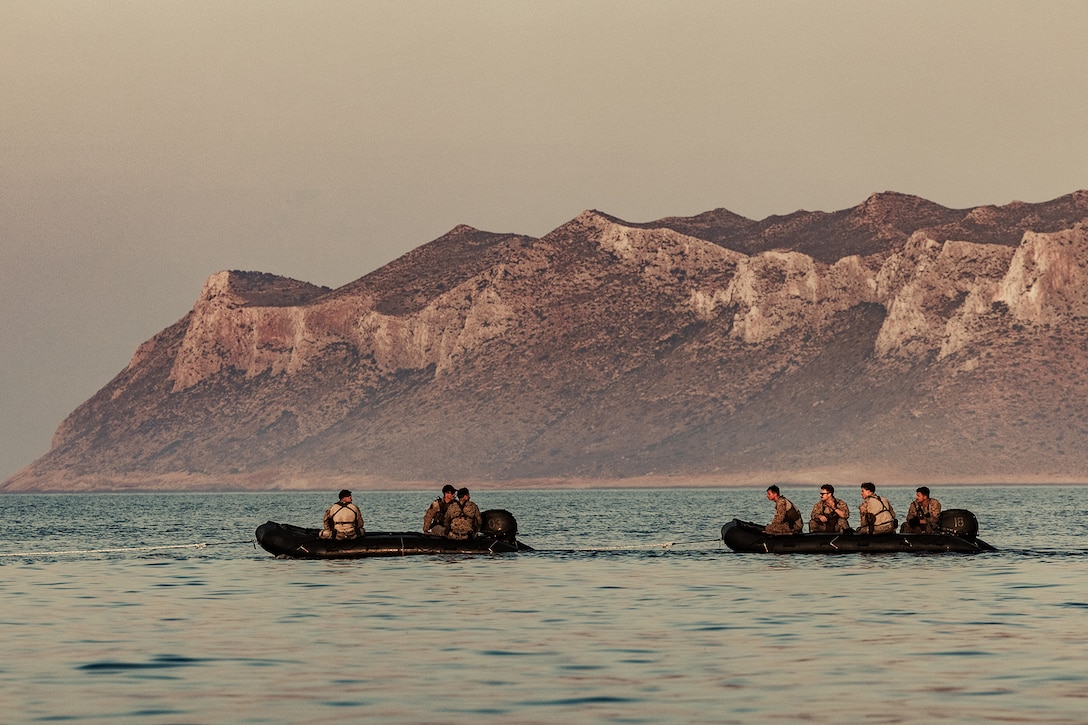Marines ride in military rafts in open water with mountains in the background.