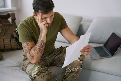 Upset member of military holding paper before himself while leaning head on his hand and reading information.