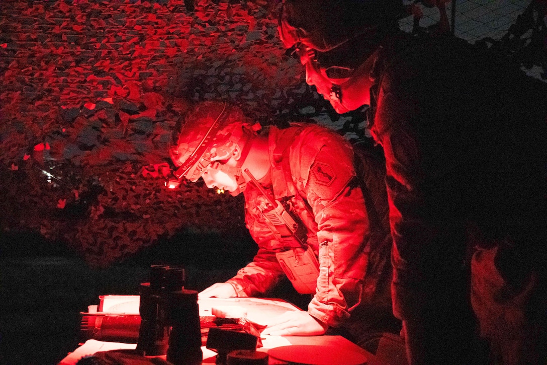 Two soldiers wearing protective gear look at paper on a table in a makeshift shelter illuminated by helmet spotlights.