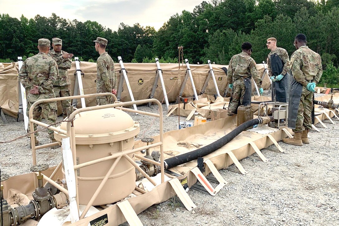 A group of uniformed service members stand by military equipment in a dirt field.