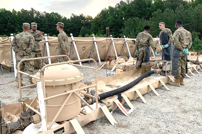 A group of uniformed service members stand by military equipment in a dirt field.