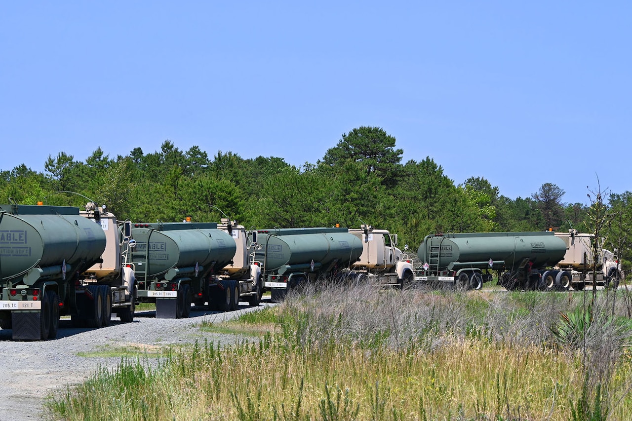 A line of four military fuel trucks sit on a gravel road.