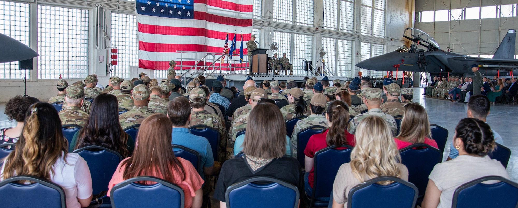 A crowd of people in a hangar listen to Maj. Gen. Michael Koscheski speech give a speech during the 366th Fighter Wing change of command ceremony, with an F-15E Strike Eagle in the background.