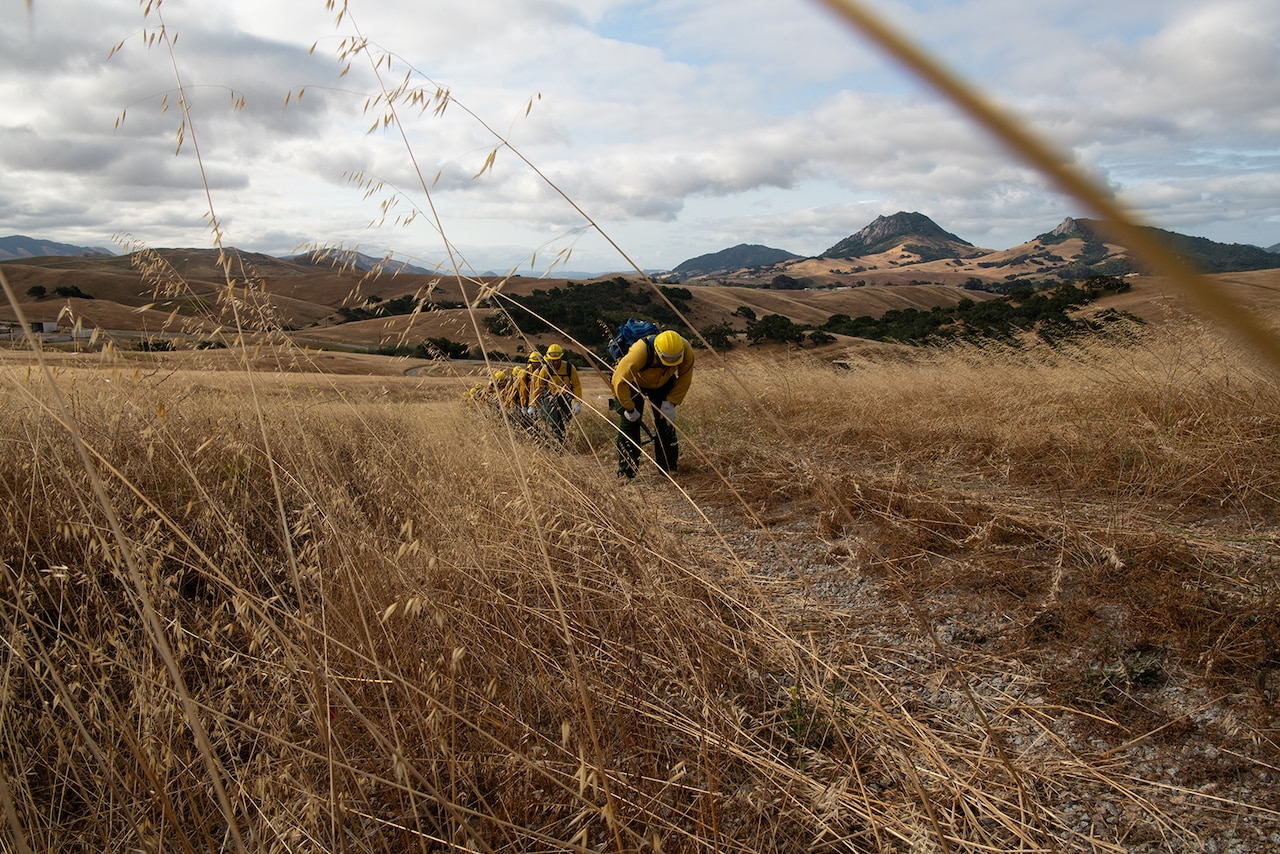 Service members wearing fire protection clothing hike up a hill with hand tools.