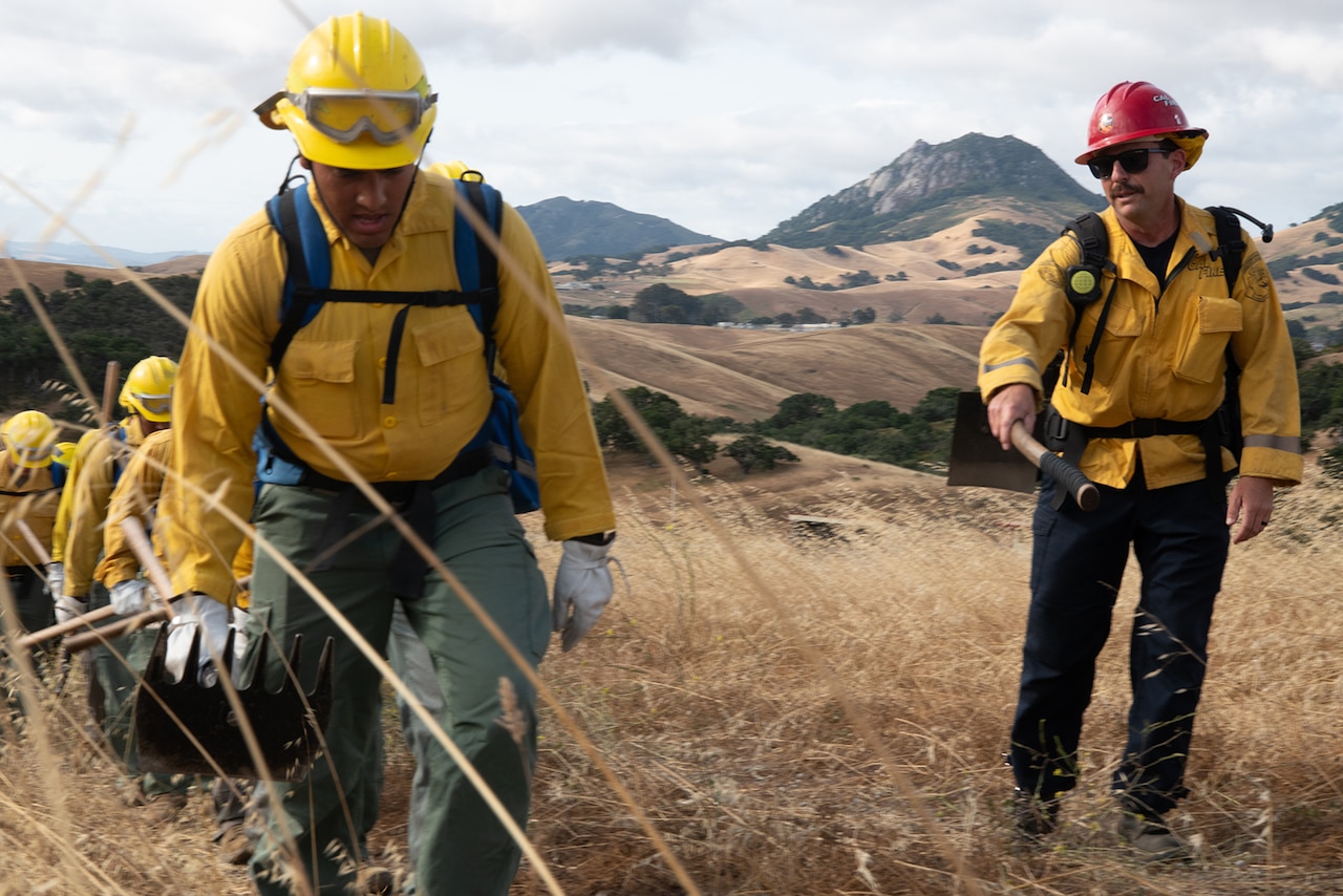 Service members wearing fire protection clothing hike up a hill with hand tools.
