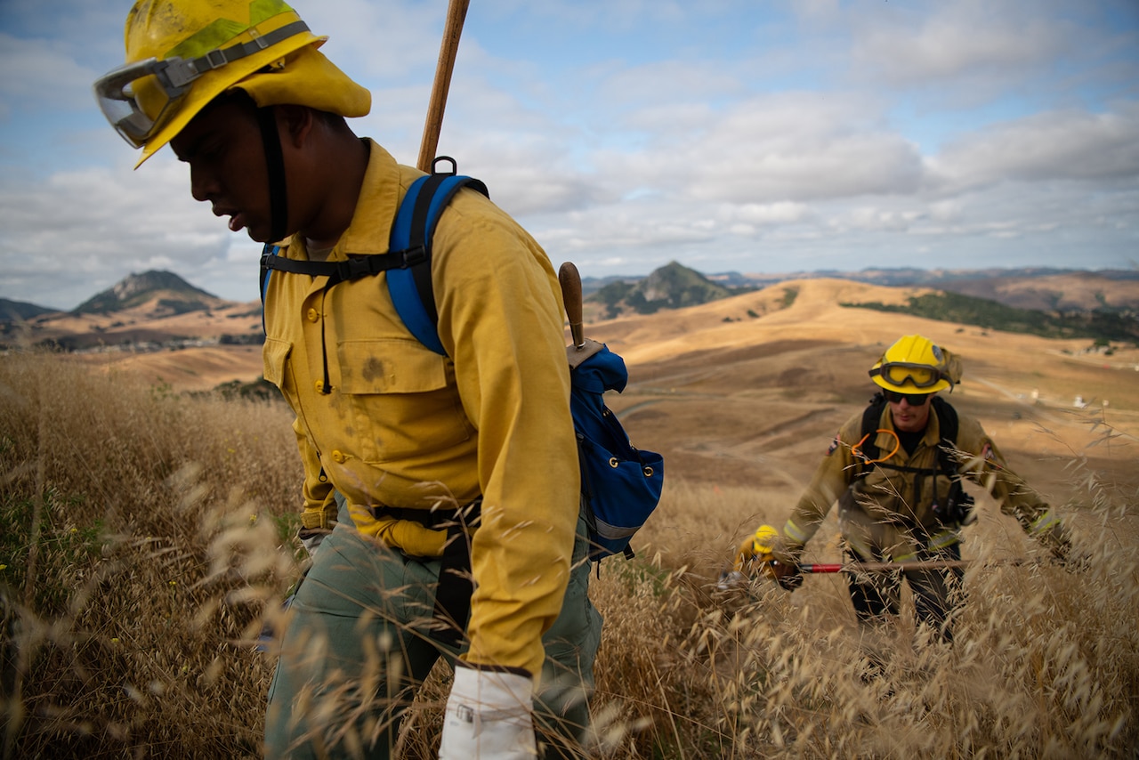 Service members wearing fire protection clothing hike up a hill carrying hand tools.