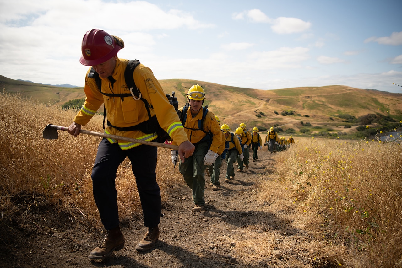 Service members wearing fire protection clothing walk in a line up a steep hill.