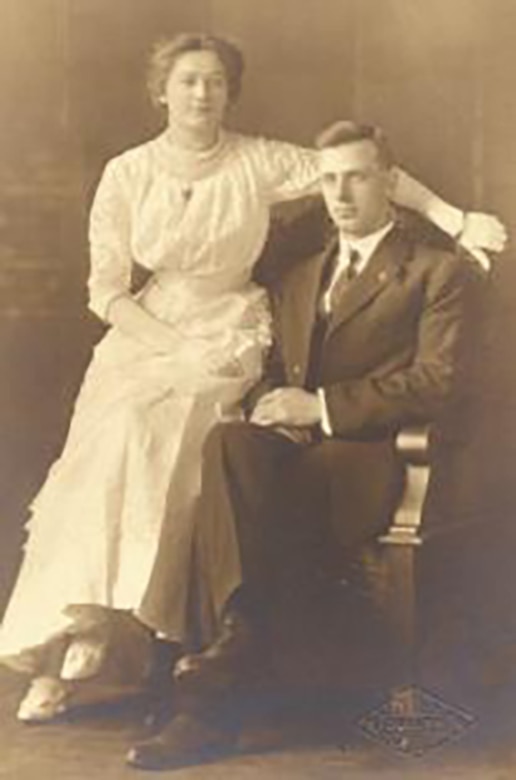 A man is seated in a chair; a woman sits on the arm rest with her left arm around the man.