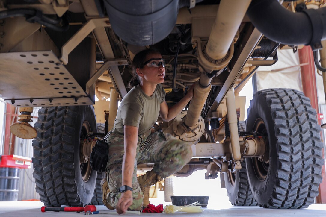 A soldier crouches under a military vehicle to perform maintenance.