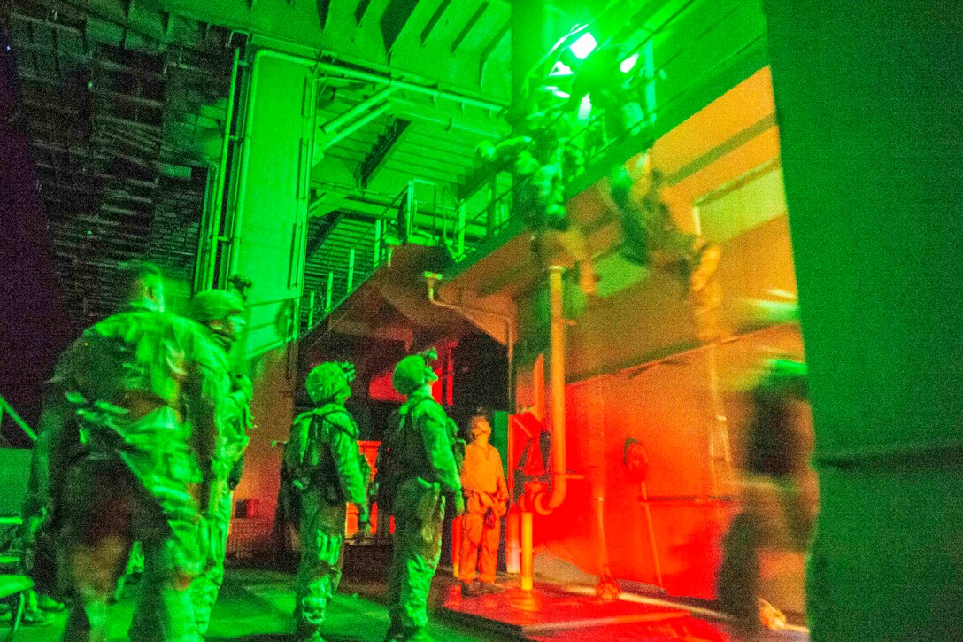 A blurry shot of two Marines in tactical gear climbing ropes attached to a balcony as fellow Marines watch illuminated by overhead lights.