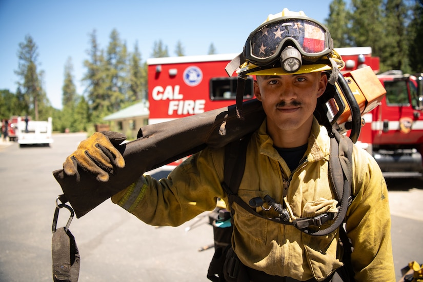 A young man wearing firefighting gear holds a chainsaw on his shoulder.