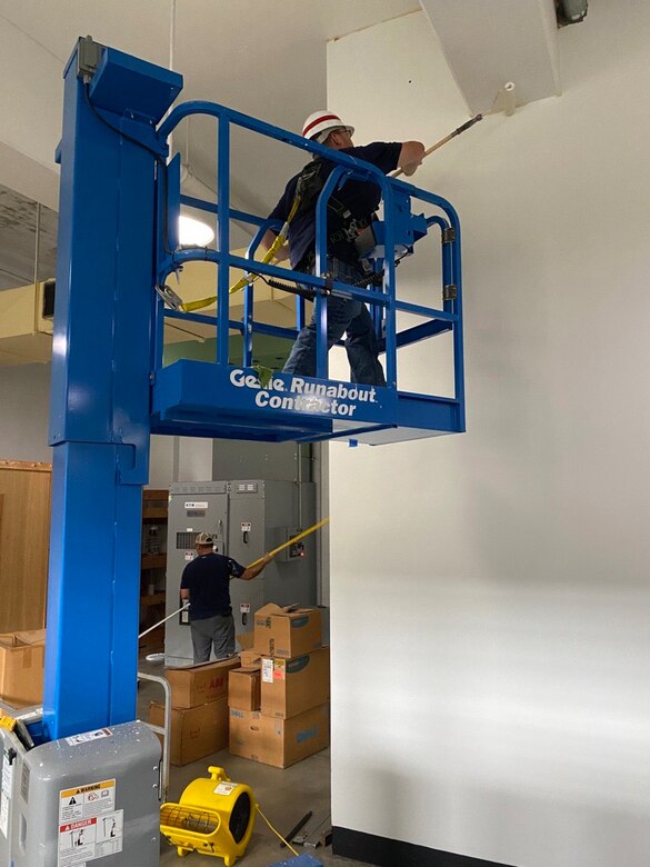 One man stands on a maintenance stand while the other stand on the ground to paint the walls.
