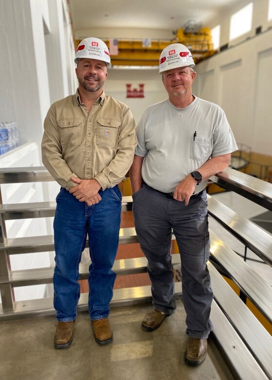 Two men stand in front of three generators.
