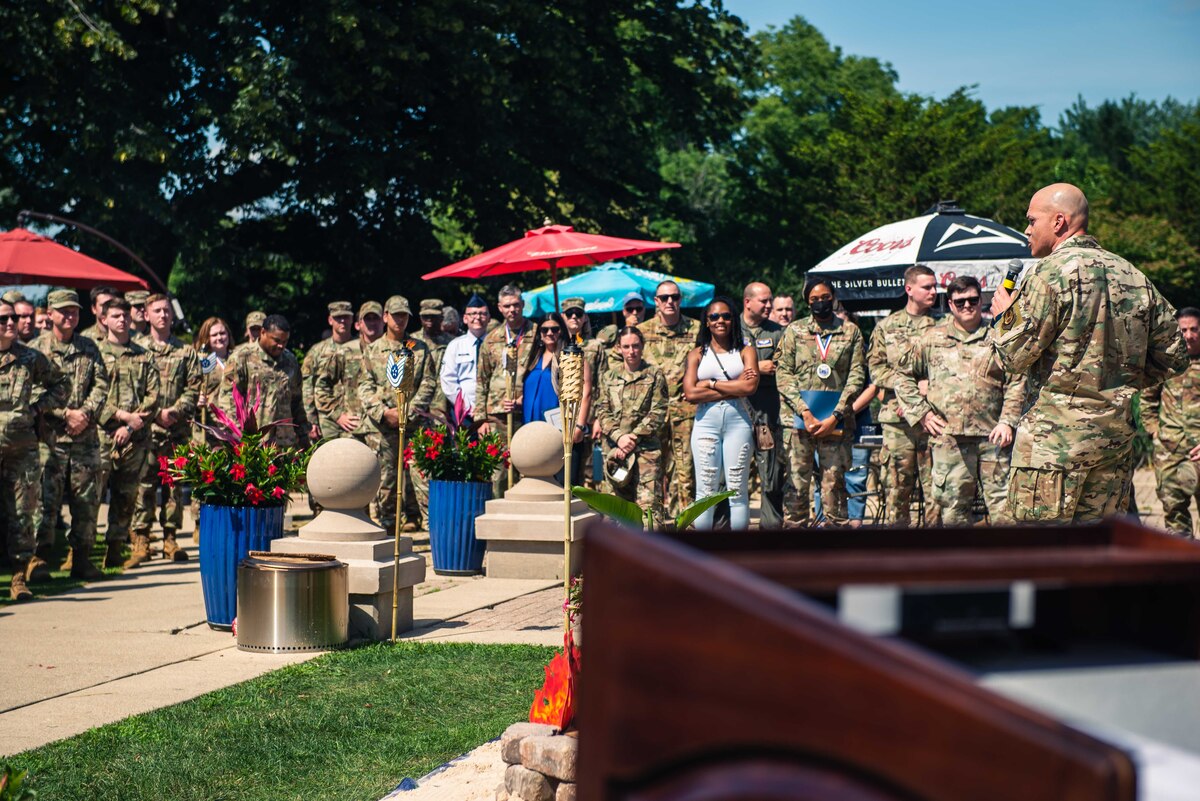 Man on right side speaking into a microphone in front of a crowd of people in uniform