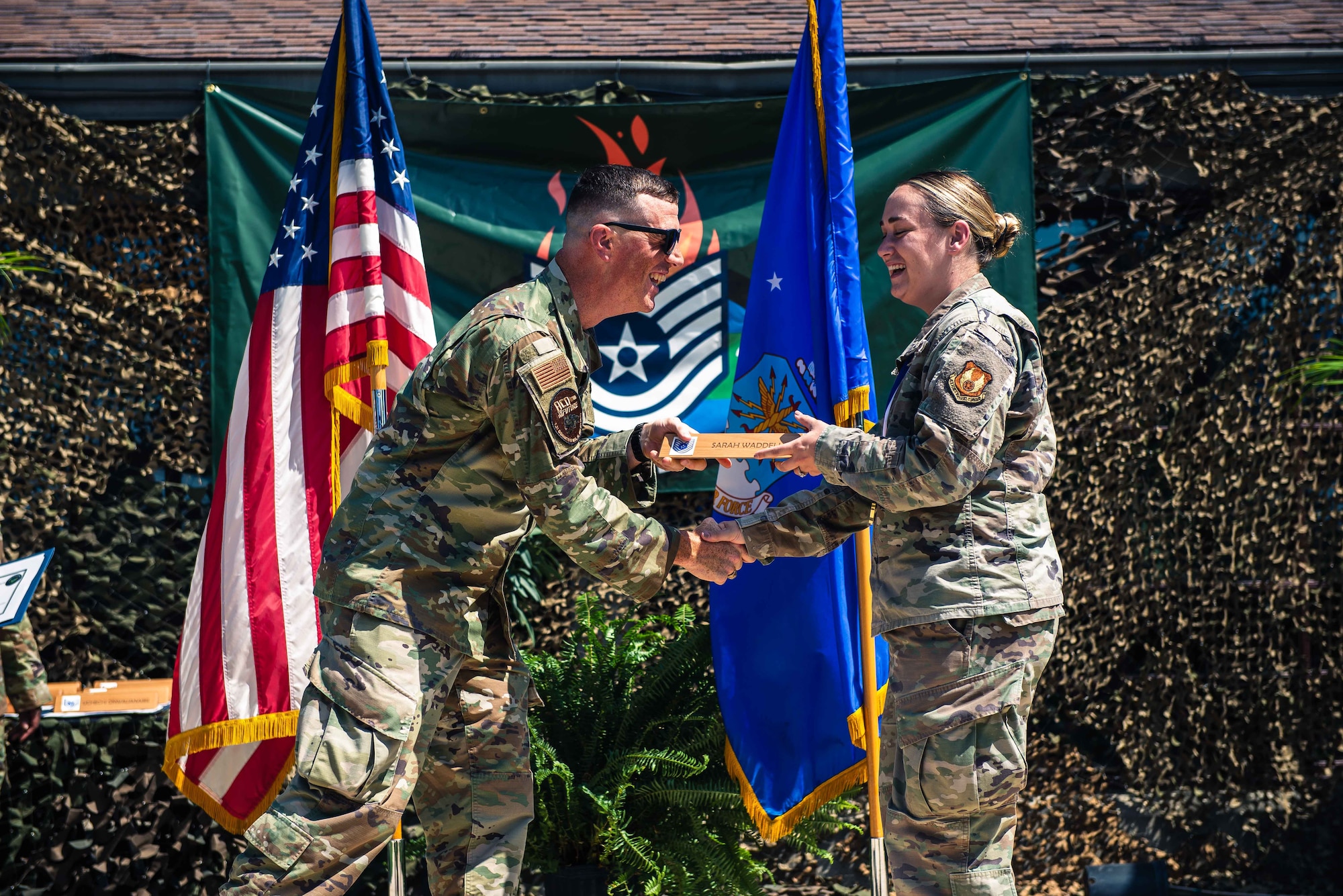 Man smiling passing a plaque to a woman in front of flags.