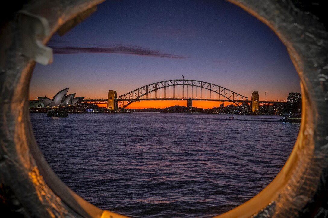 Ships sail in a body of water near a bridge with buildings in the background under an orangish sky as seen through a ship’s porthole.
