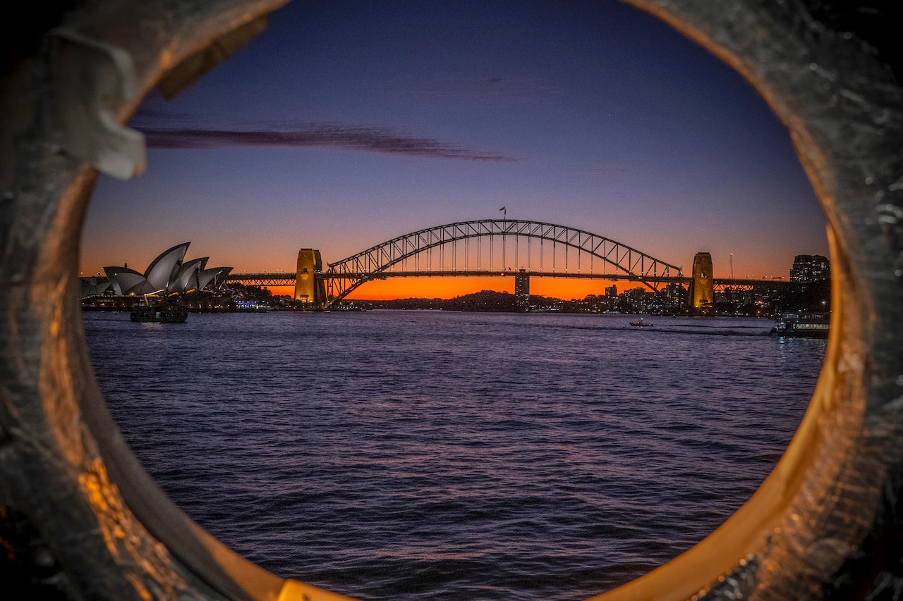 Ships sail in a body of water near a bridge with buildings in the background under an orangish sky as seen through a ship’s porthole.