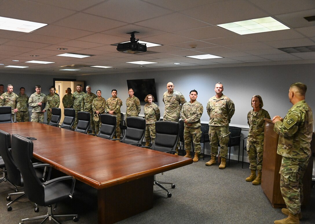 A group of military members in uniform listen to a person speaking.