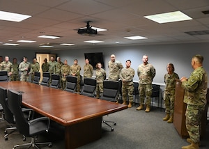 A group of military members in uniform listen to a person speaking.