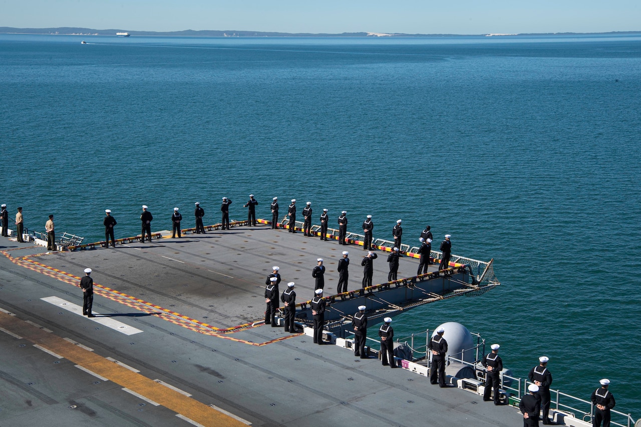 Sailors and Marines in uniform stand at the ship’s perimeter.
