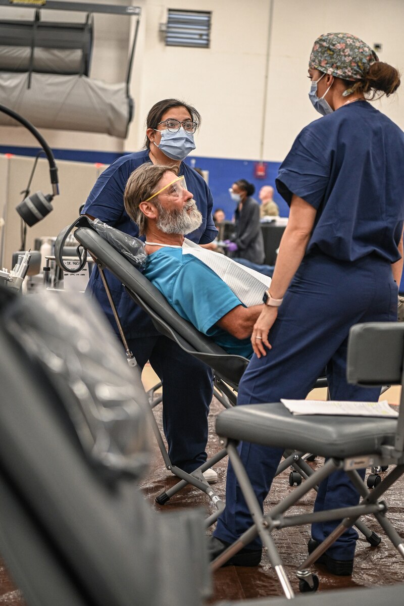 two dental workers talk with a patient sitting in a reclining dental chair.