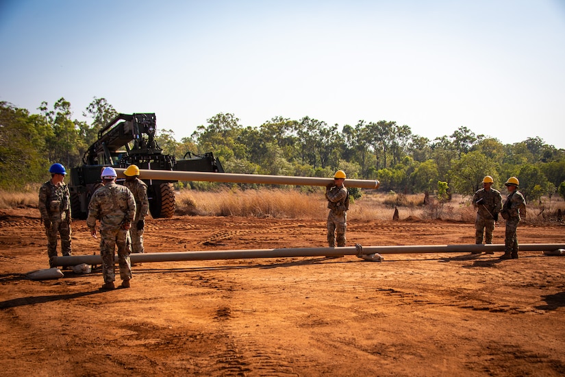 Soldiers wearing hard hats carry a large pipe.