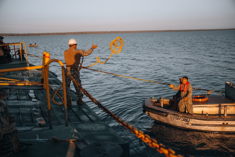 A sailor throws a rope from a large vessel to sailors on a smaller boat.