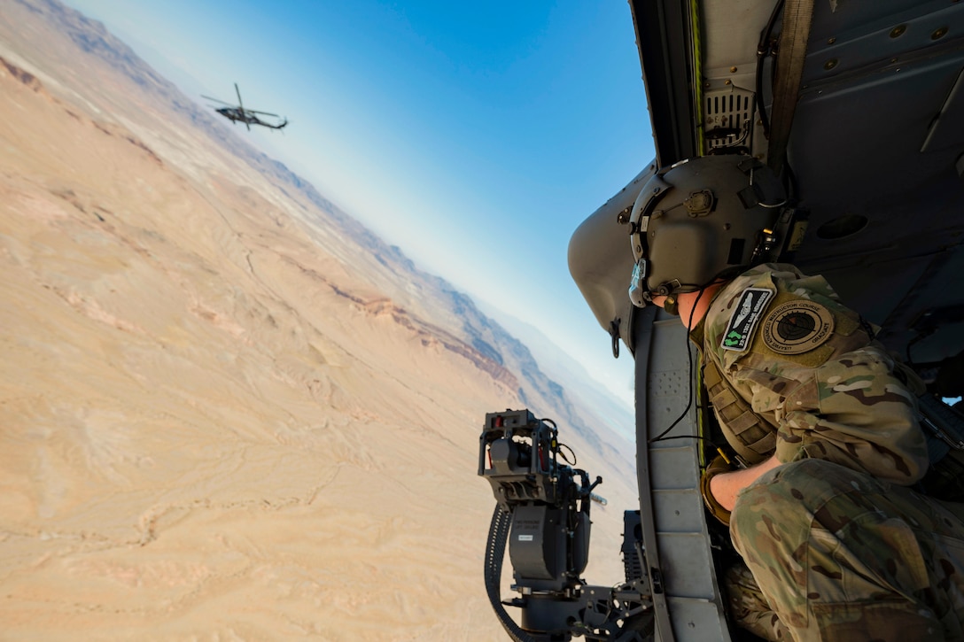 An airman wearing protective gear looks out the open doorway of an airborne helicopter as another flies by over a desert-like area.