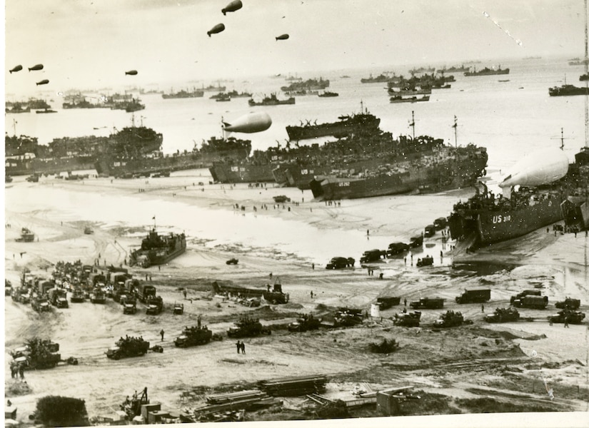 Several ships rest on a beachhead as troops and dozens of small vehicles move around on the sand.