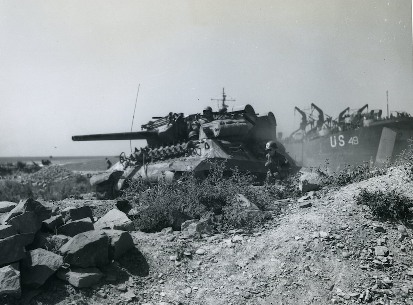 A tank rolls along rocky ground as a ship sits off the waters in the background.