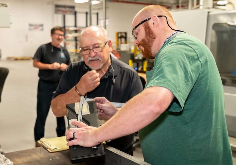 The image depicts two people examining a piece of equipment on a work table in an industrial work area.