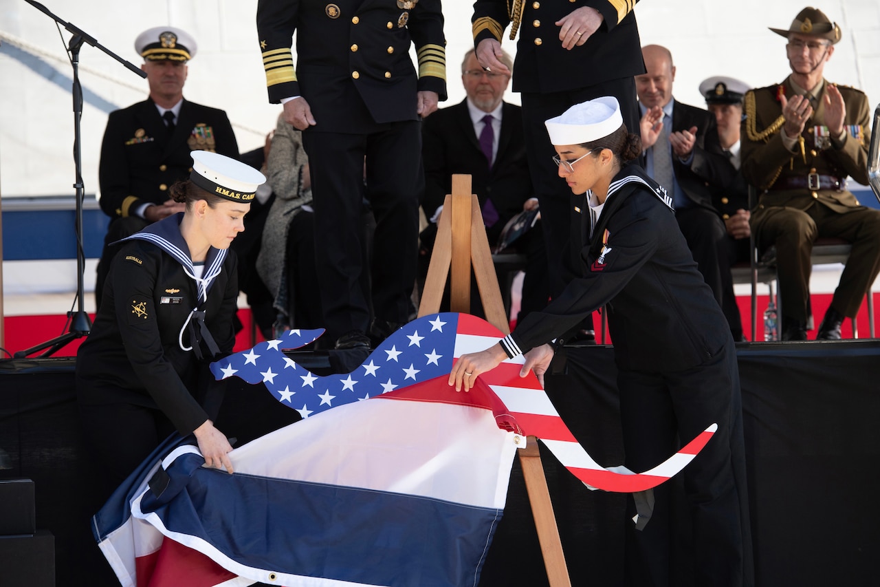 A U.S. sailor and an Australian sailor unveil and a kangaroo emblem with a U.S. flag design in front of an applauding audience.