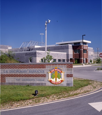 A red and grey brick building is in the background with a red brick sign in the foreground. Grass is in the foreground and blue sky in the background.