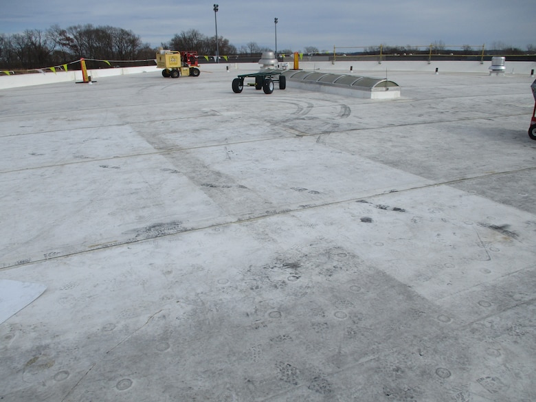 Grey roofing material is in the foreground with construction equipment, trees and the sky in the background.