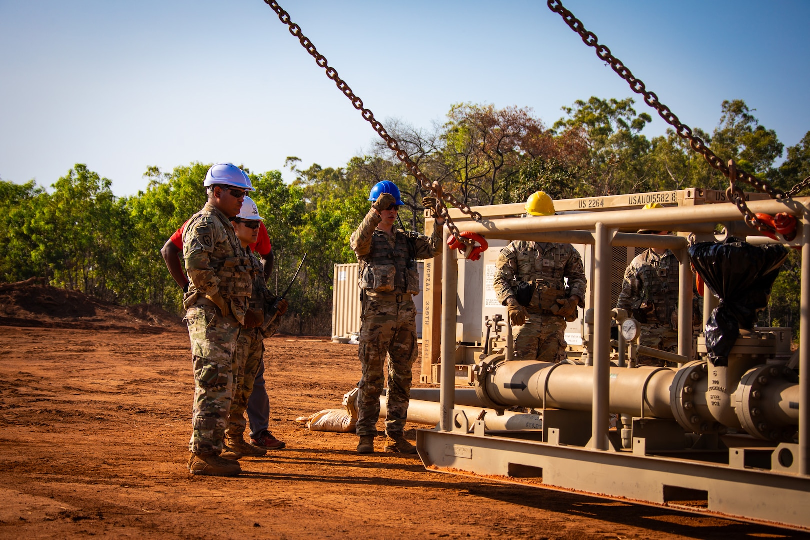 Soldiers wearing hard hats stand near a large pump suspended by chains.