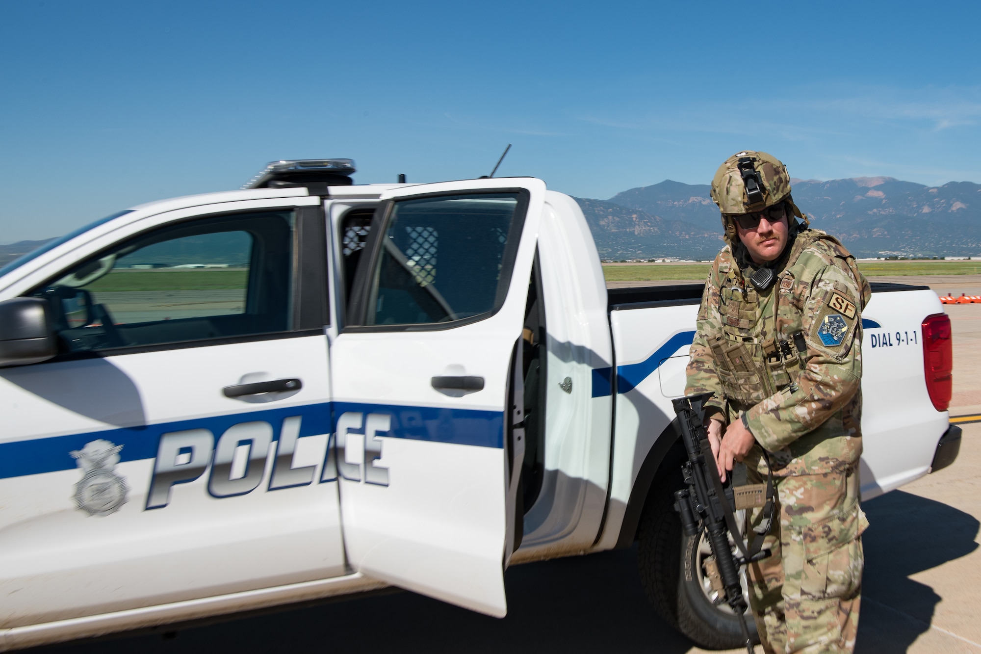 An Airman armed with a rifle stands next to a police truck on the flight line with the Rocky Mountains in the background.