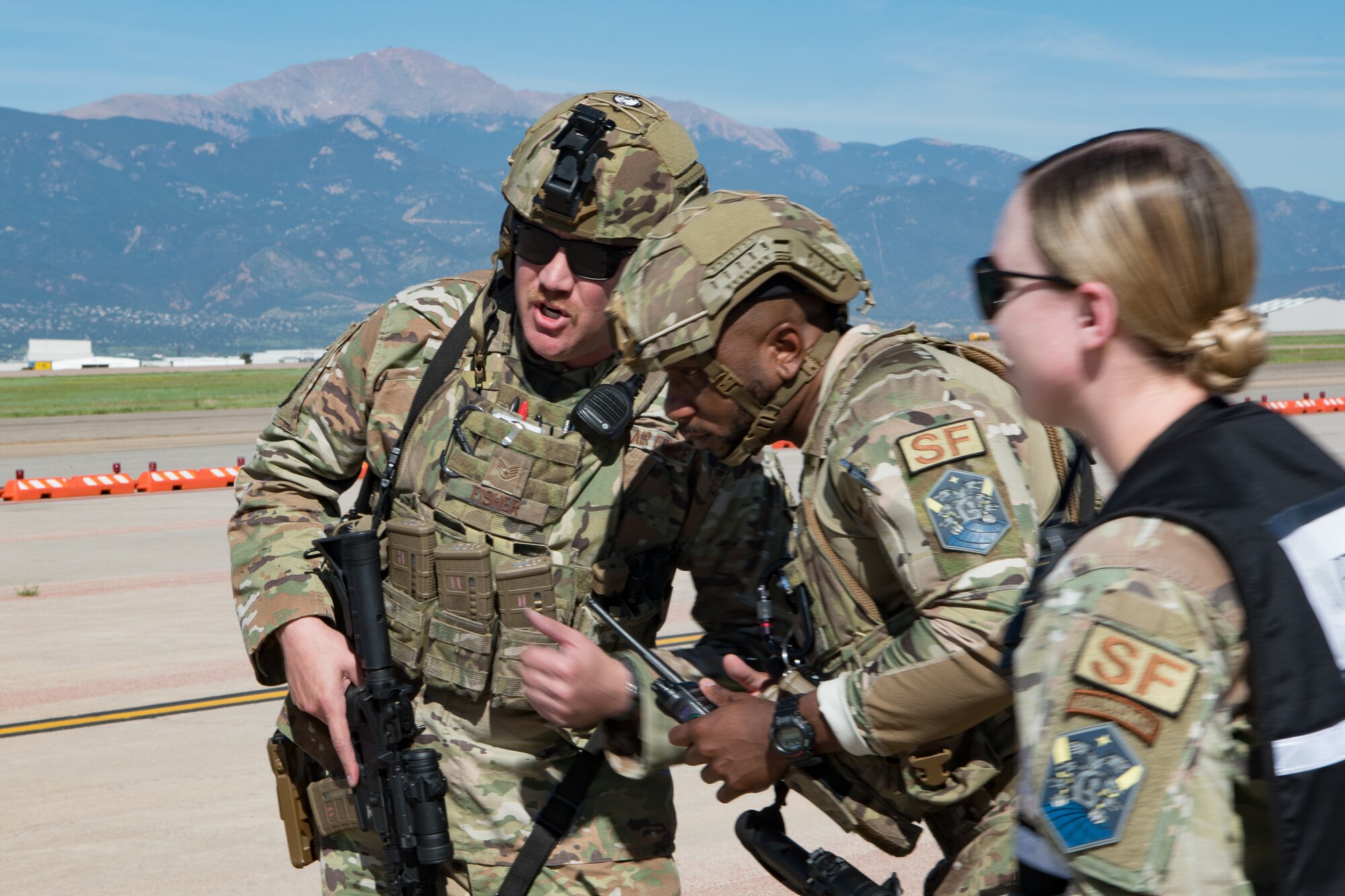 Two Airmen speak to each other on a flight line while another Airman wearing a vest observes with the Rocky Mountains in the background.