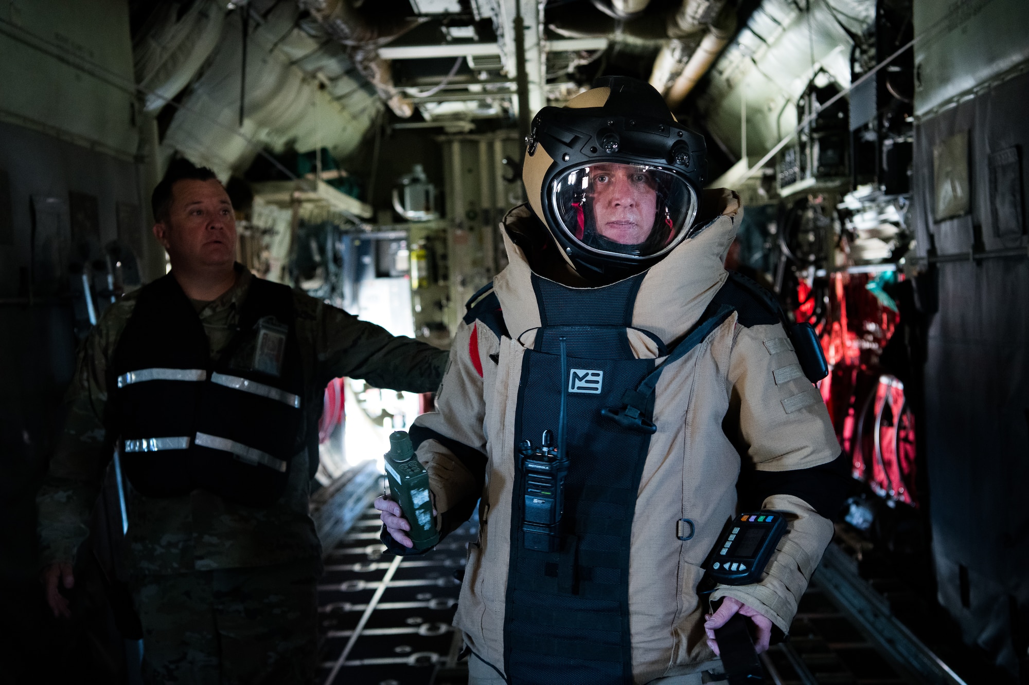 An Airman wearing an explosive ordnance disposal protective suit holds a handheld piece of equipment in the back of a C-130 aircraft cargo bay while a uniformed Airmen in a vest observes in the background.
