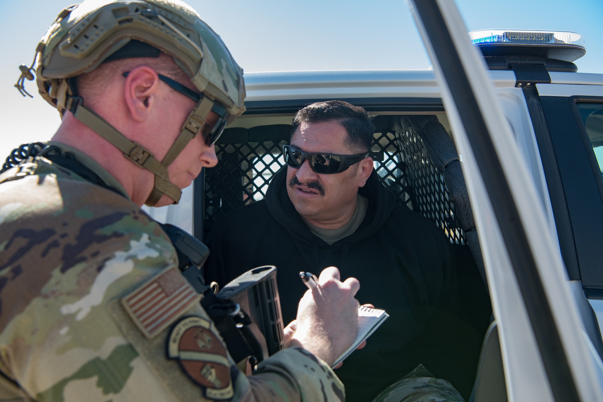 A uniformed Airman writing on a notepad while speaking to an Airman wearing a black sweatshirt sitting in the rear seat of a police vehicle.