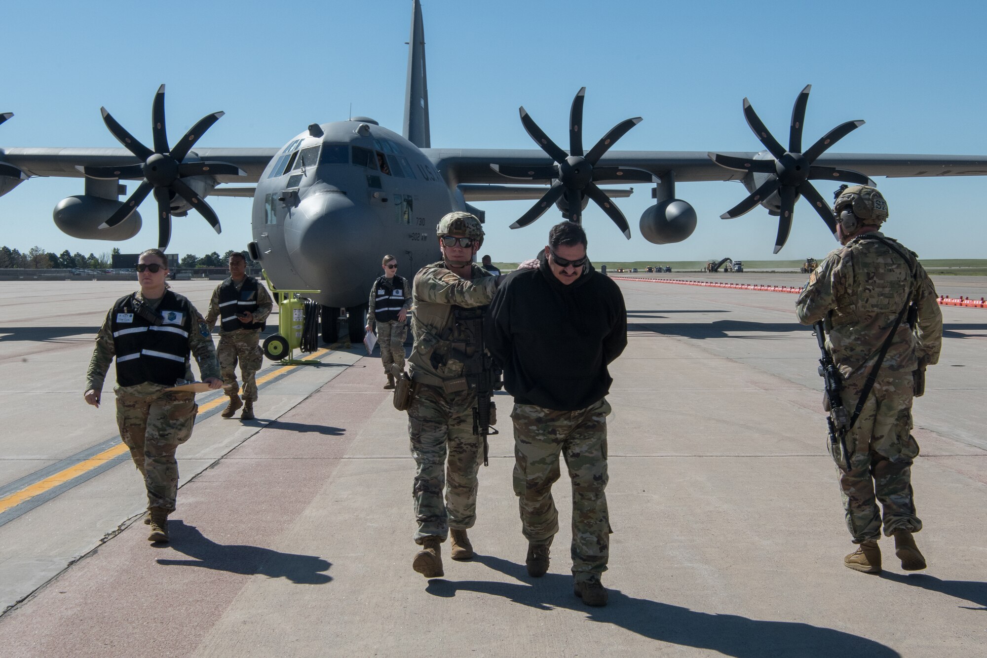 Five Airmen walking away from a C-130 aircraft escorting an additional handcuffed Airman wearing a black sweatshirt.