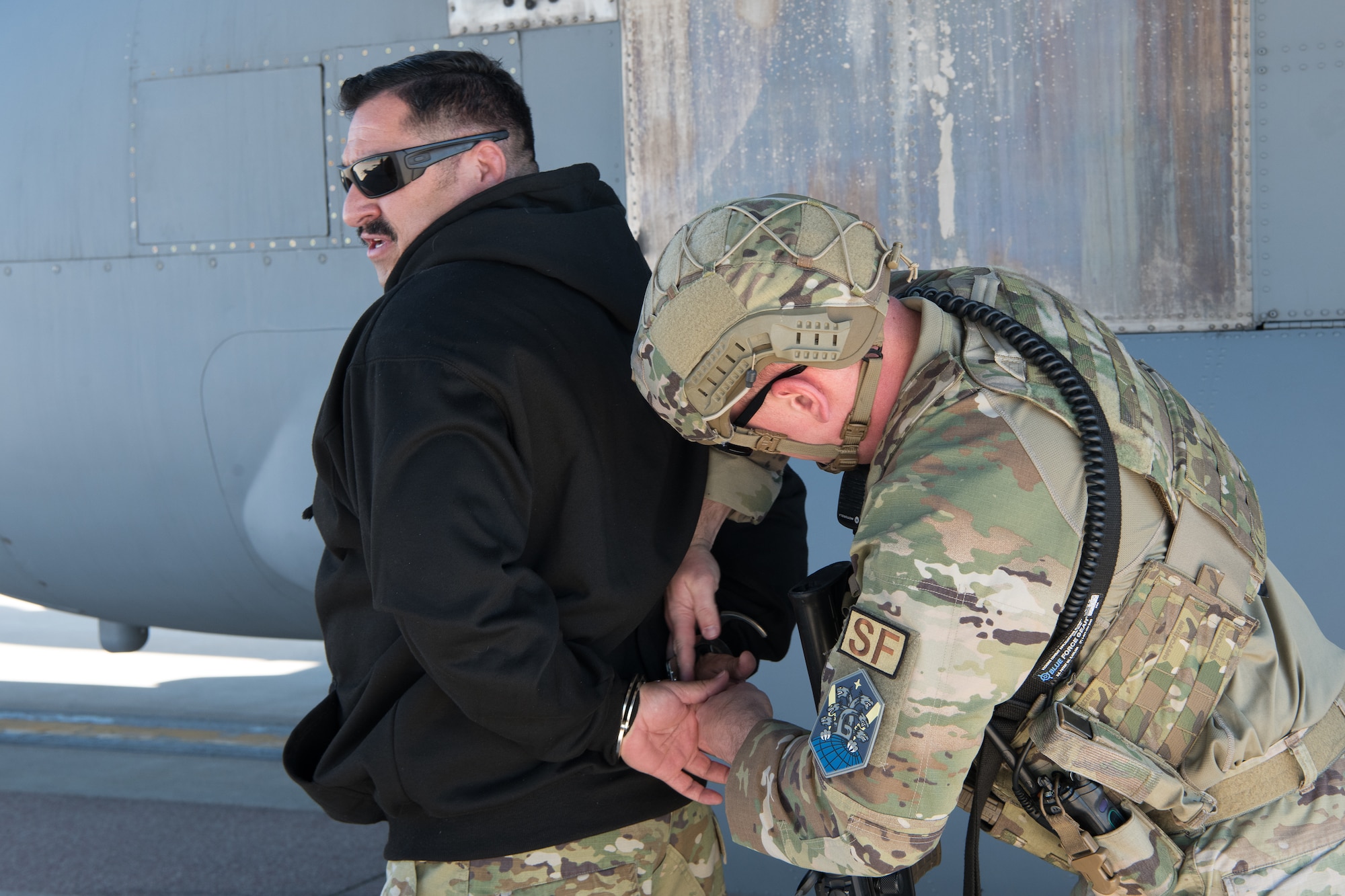 A uniformed Airman handcuffing a man wearing a black sweatshirt in front of a C-130 aircraft.