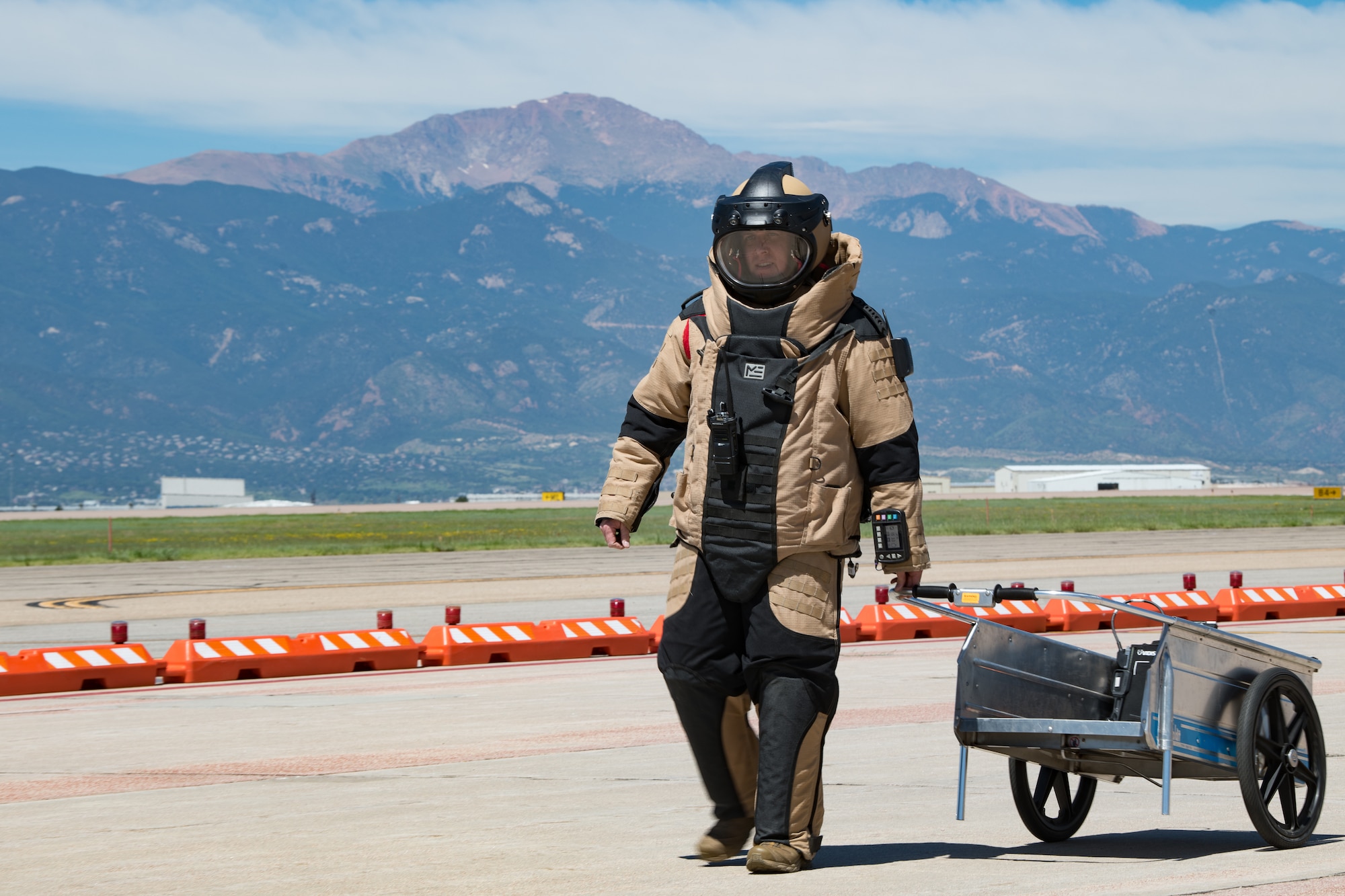 An Airman wearing an explosive ordnance disposal protective outfit pulling a cart behind him on a flight line with Pikes Peak in the background.