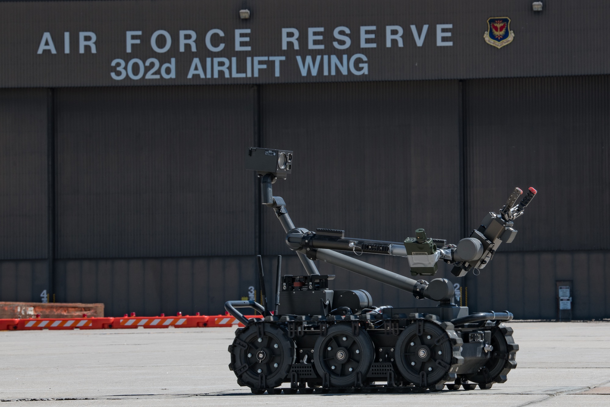 A small remote-controlled robot traveling along the flight line propelled by a continuous track in front of a hangar labeled "Air Force Reserve, 302nd Airlift Wing."