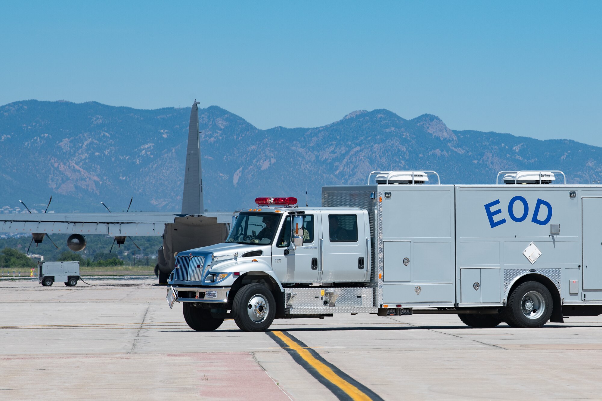 A truck with a compartment attached to the rear labeled EOD on a flight line with a C-130 aircraft behind it and mountains in the distance.
